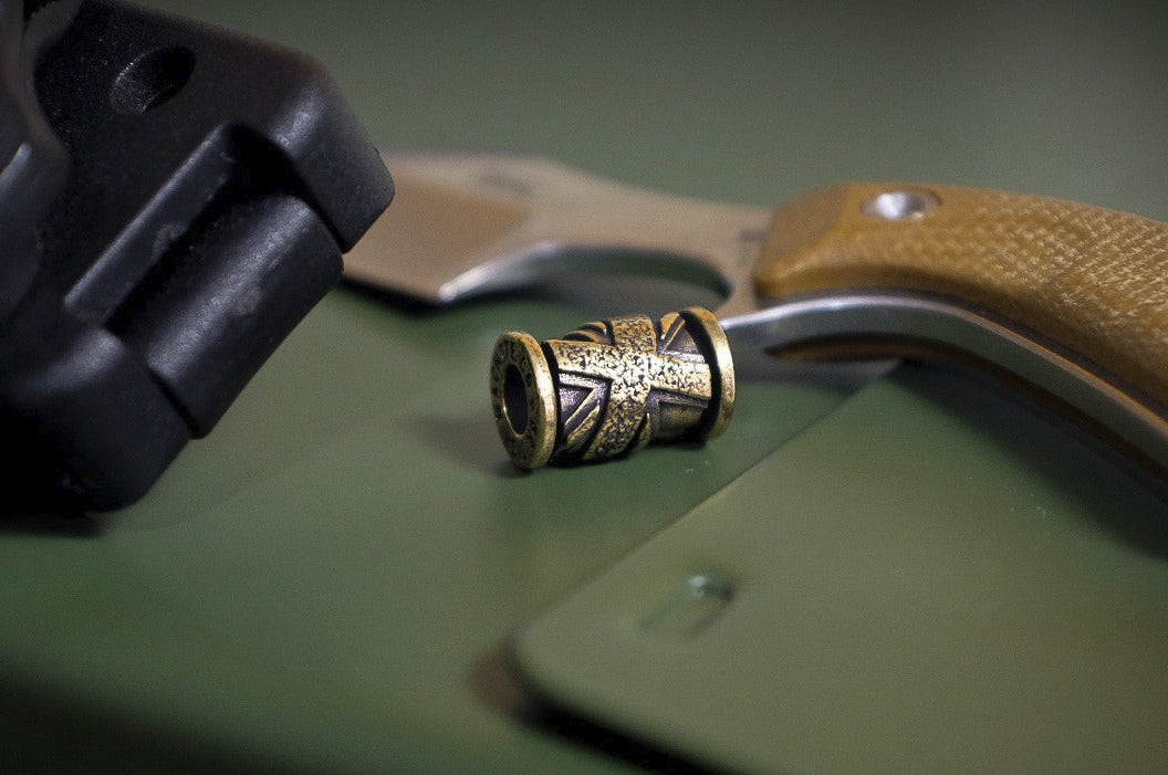 Close-up of a bronze Union Jack bead on a green surface with a sheath and knife in the background