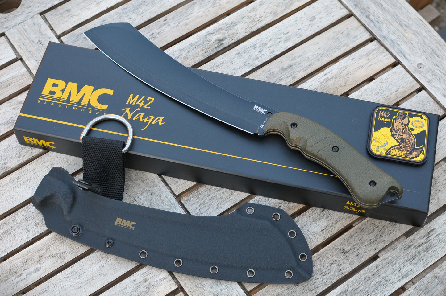 BMC M42 Naga parang with an olive green handle, shown with its black branded box, black sheath, and a small patch on a weathered wooden slat surface.