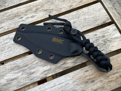 A black Kydex knife sheath with a black paracord-wrapped handle and a gold BMC logo, displayed on a weathered wooden slat surface.