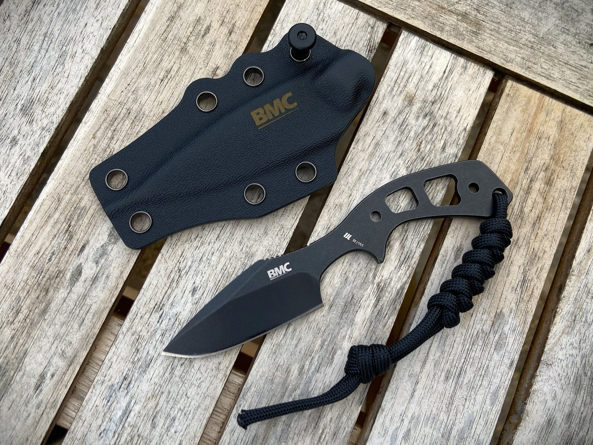 A black knife with a paracord lanyard and a matching black BMC sheath, displayed on a weathered wooden plank surface.
