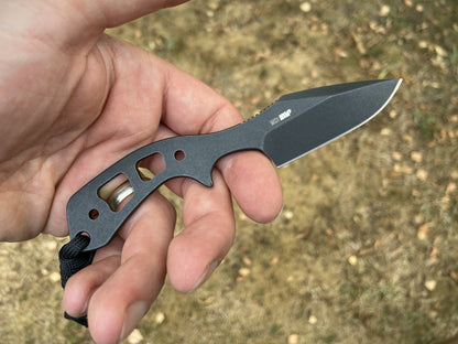 A hand holding a compact black tactical folding knife with a black blade and textured handle, set against a blurred outdoor background of dry grass and soil.