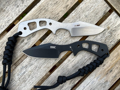Two fixed-blade knives with paracord lanyards, one with a silver handle and one with a black handle, displayed on a weathered wooden slatted surface. The top knife is labeled IMP and the bottom knife is labeled BMC.