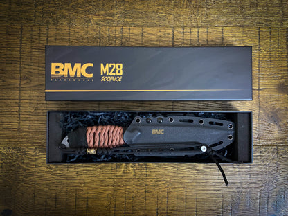 BMC M28 custom blade shown in its open presentation box on a wooden surface, with the branded lid placed above. The product includes a black metal bladeskate with a copper braided handle wrap and black grip, nestled in dark blue paper filler.