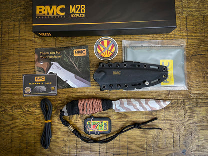 BMC M28 Solifuge knife set laid out on a wooden surface, including the black box, sheath, knife with patterned handle, lanyard, warranty card, and other included accessories.