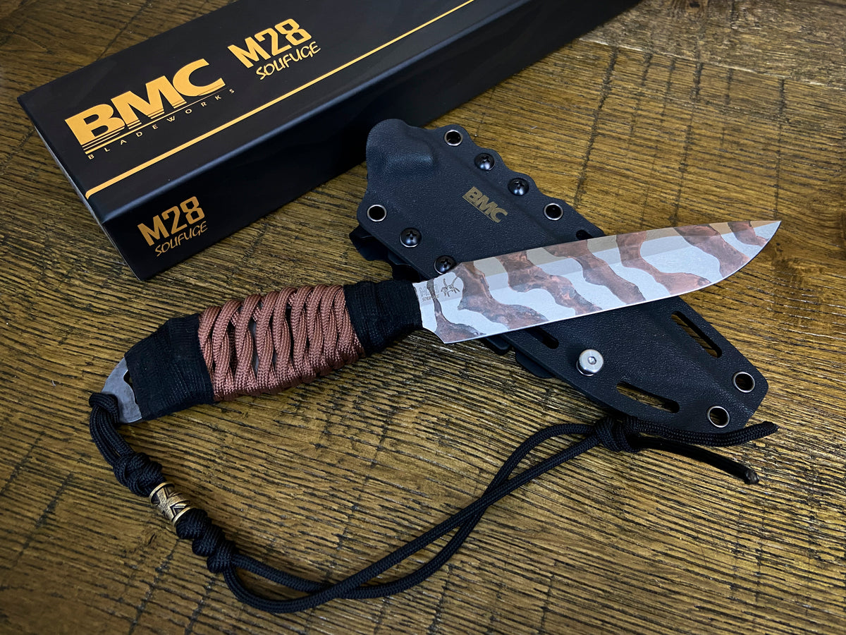 BMC M28 Solifuge fixed blade knife with a tiger-stripe pattern blade, wrapped handle, and black sheath, displayed on a wooden surface next to its black branded box.