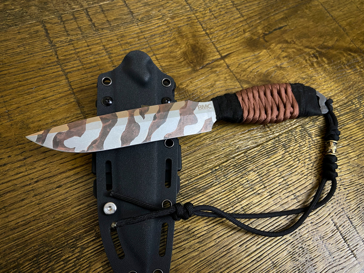 A fixed-blade knife with a brown, beige, and white camouflage patterned blade, wrapped in brown cord near the handle, shown resting on its black sheath over a wooden surface.