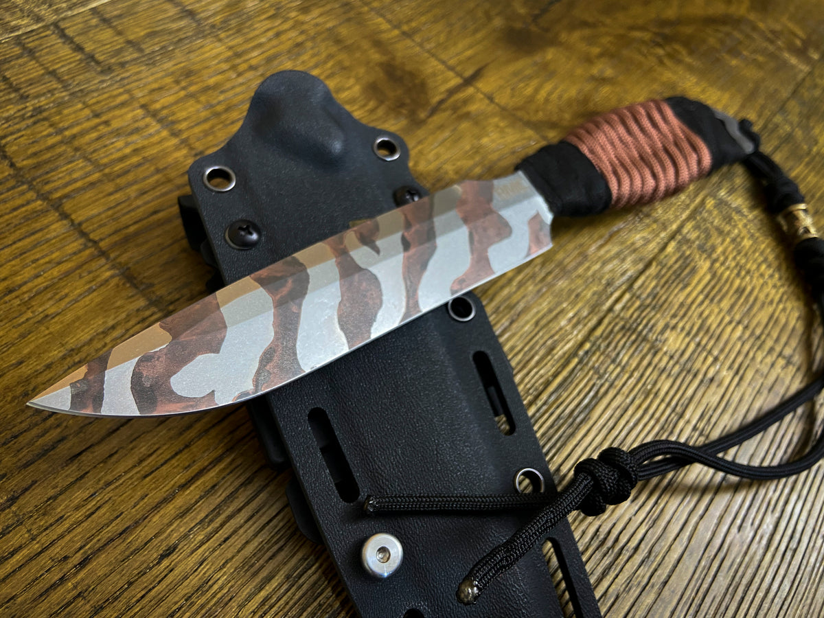 A fixed-blade knife with a brown, beige, and gray camouflage pattern blade, a black wrapped handle with a copper-colored wrap, and a black lanyard, resting on its black sheath over a wooden surface.