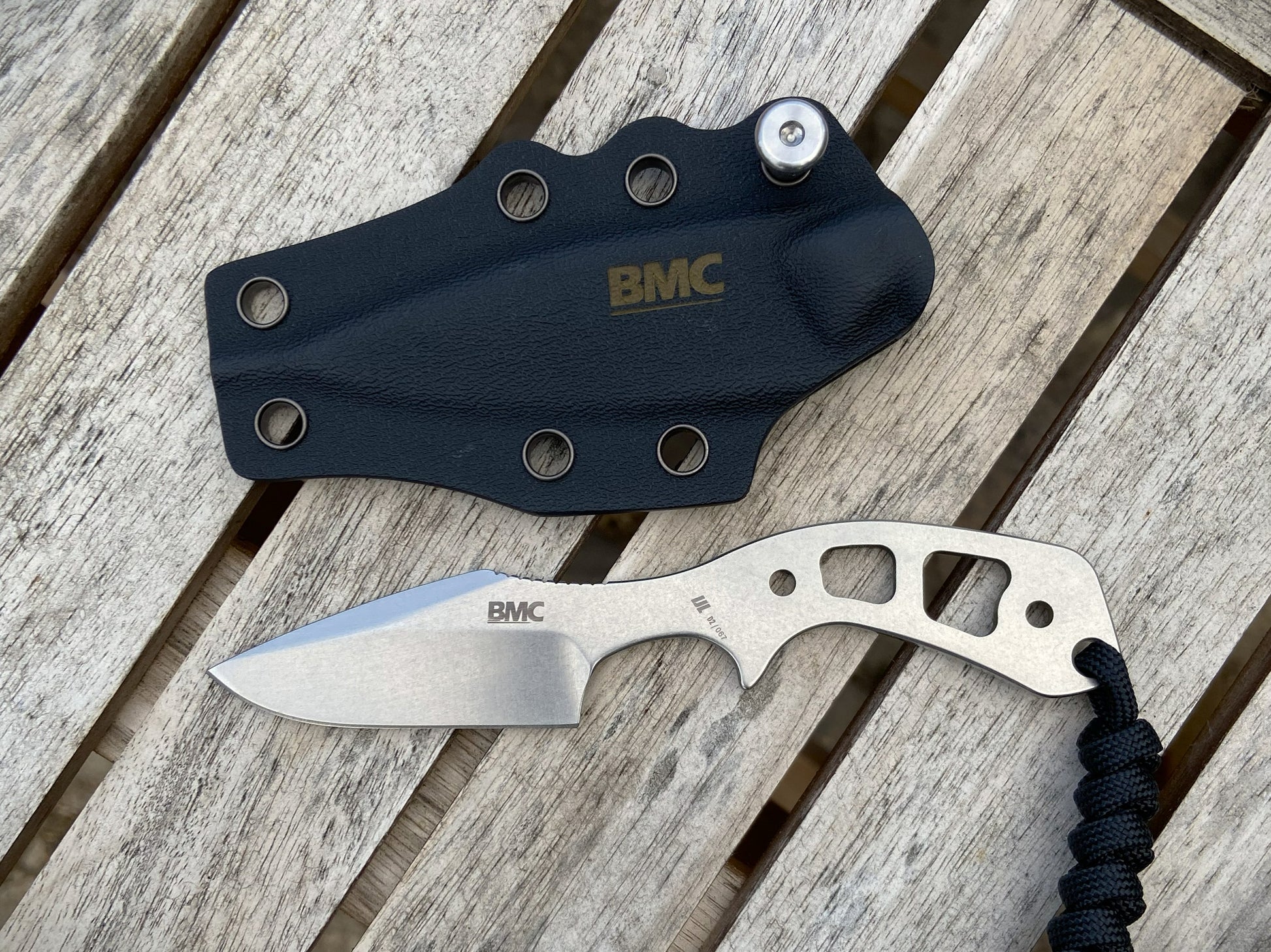 A BMC fixed-blade knife with a silver handle and black paracord lanyard, displayed next to its black Kydex sheath with gold BMC branding, set on a weathered wooden plank surface.