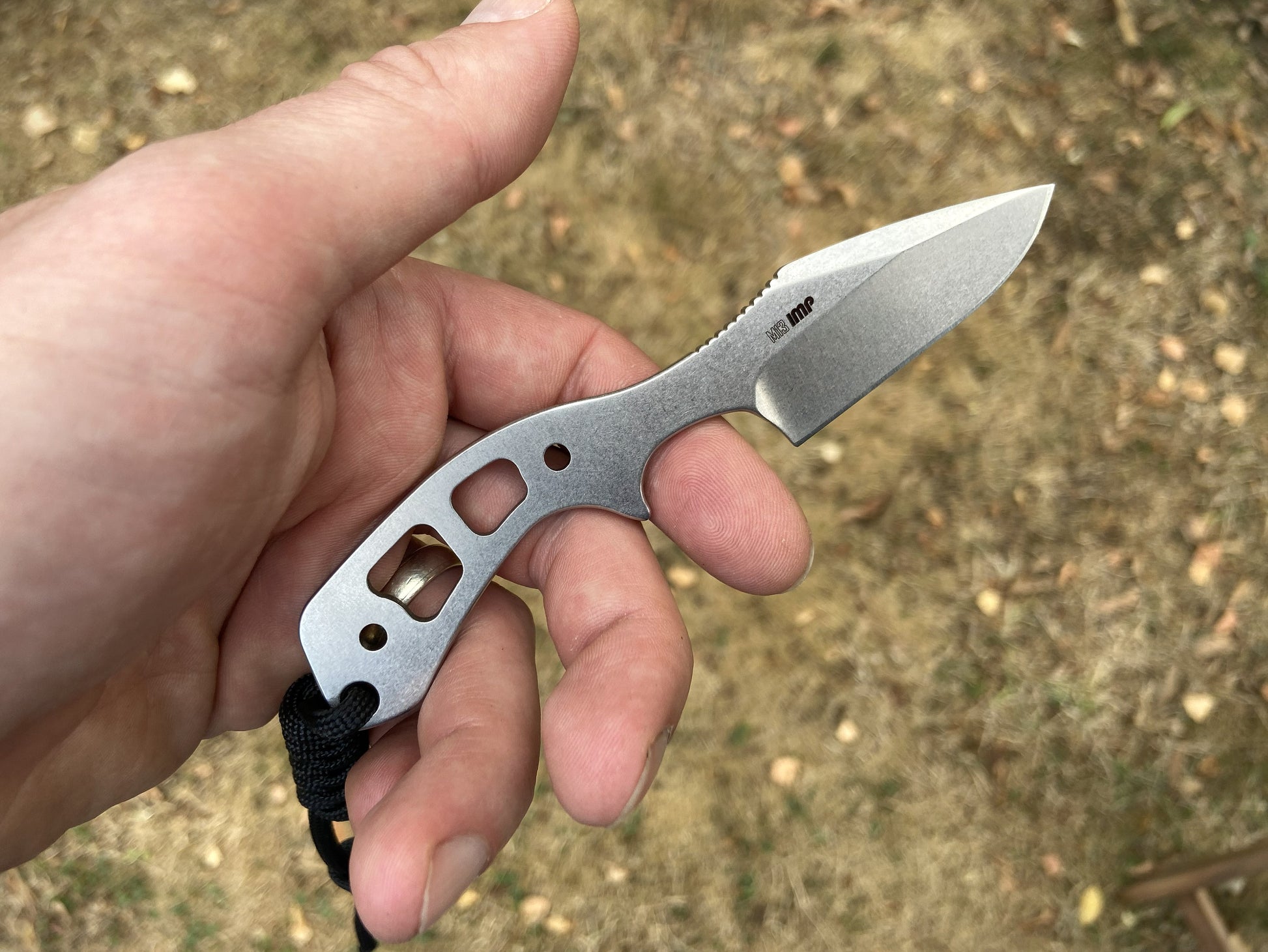 A hand holding a small fixed-blade knife with a silver metal handle and blade, photographed outdoors against a blurred ground background.