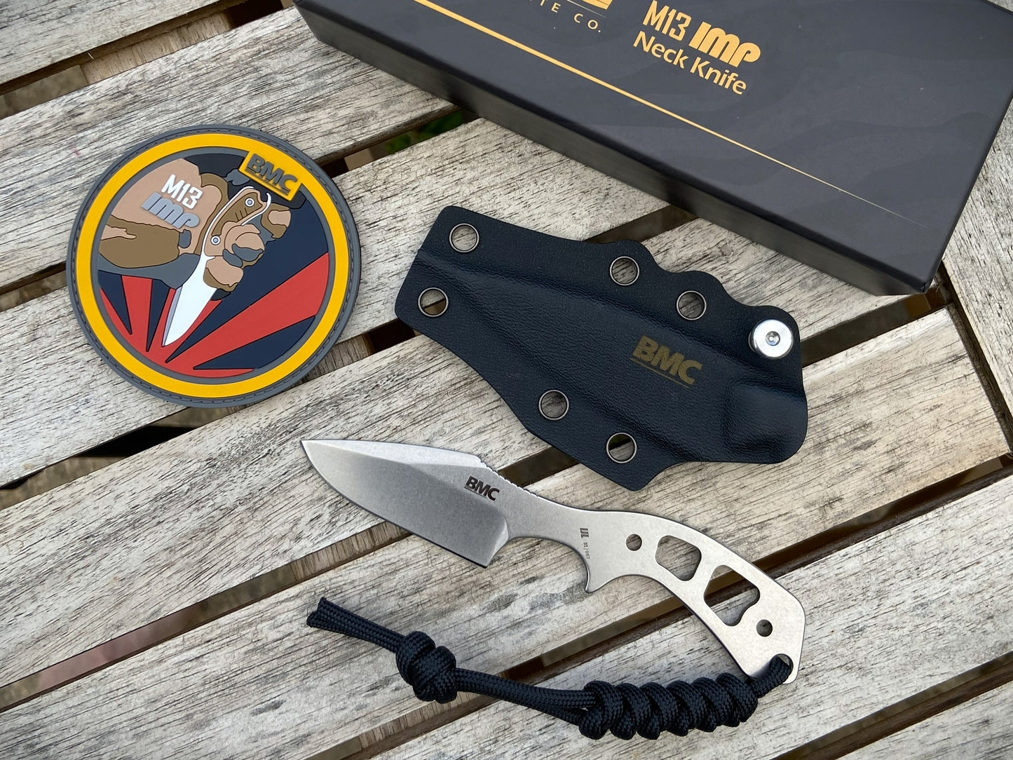 A BMC M13 neck knife with a silver blade and handle, displayed on a weathered wooden surface alongside its black sheath, a circular patch, and the branded box.