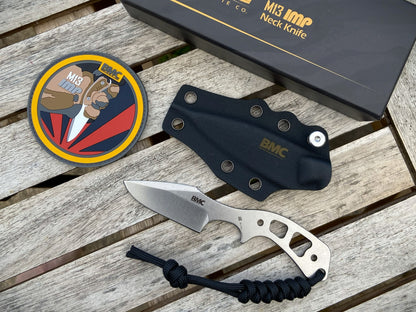 A BMC M13 neck knife with a silver blade and handle, displayed on a weathered wooden surface alongside its black sheath, a circular patch, and the branded box.