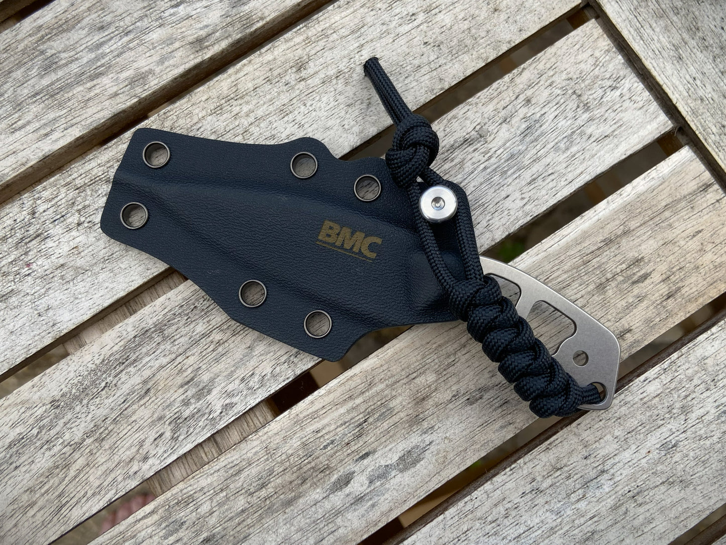 A black BMC Kydex sheath holding a BMC knife with a black paracord lanyard, placed on a weathered gray wooden slatted surface.